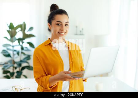 Ritratto di una giovane donna indiana o araba, manager, copywriter creativo, hr, che tiene in mano un notebook aperto mentre si trova in un ufficio moderno, guarda la fotocamera e sorride Foto Stock