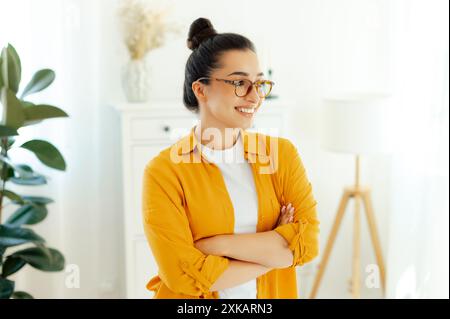 Elegante giovane donna di etnia araba o indiana con una camicia arancione con occhiali da vista, dipendente dell'azienda, direttore creativo si trova all'interno vicino alla scrivania con le braccia incrociate, guarda lontano e sorride Foto Stock
