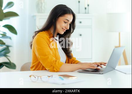 Una giovane donna araba o indiana, esperta di IT, product manager, web designer, lavora in un luogo di lavoro con un notebook su un progetto, guarda lo schermo di un notebook e sorride Foto Stock