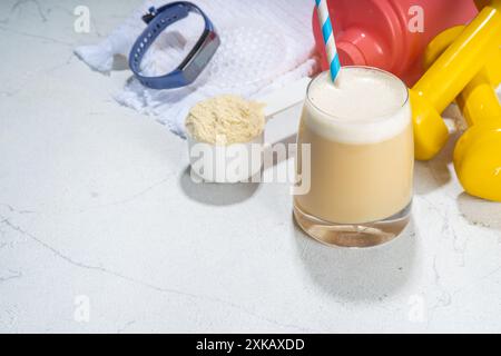 Vetro bianco vaniglia per agitare le proteine, con cannuccia, con polvere proteica, manubri, asciugamano e accessori fitness, su copia sfondo bianco del tavolo sport bar Foto Stock