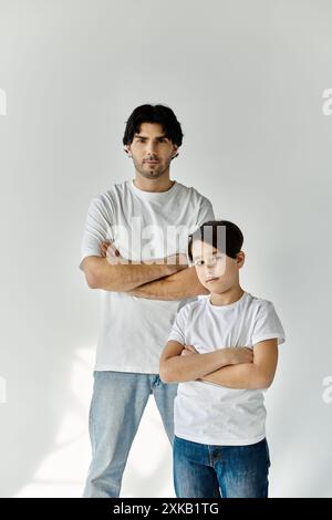 Un padre e suo figlio stanno fianco a fianco, entrambi indossando t-shirt bianche e jeans, con le braccia incrociate. Foto Stock