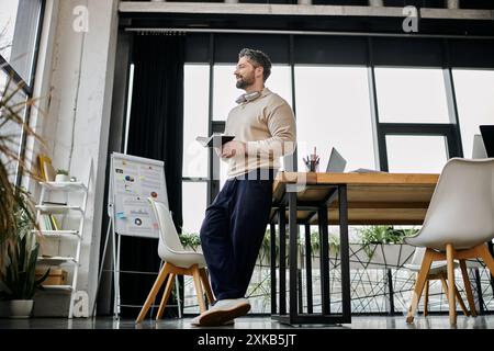 Un bell'uomo d'affari con la barba si trova in un ufficio moderno, tenendo un notebook, pensando alla sua prossima mossa. Foto Stock