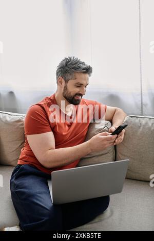 Un uomo barbuto siede su un divano in casa sua, lavorando su un portatile mentre controlla il telefono. Foto Stock