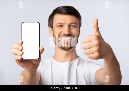 Uomo sorridente Holding Blank Cellphone gesticolando Thumbs up, isolato Foto Stock