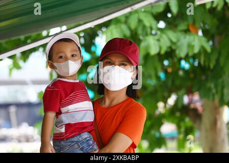 Una donna asiatica con i capelli corti e una camicia arancione sta tenendo suo figlio che indossa una camicia rossa e indossa una maschera facciale. Foto Stock