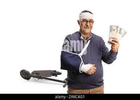 Uomo maturo sorridente con un braccio rotto e una benda sulla testa che regge pile di denaro davanti a uno scooter elettrico isolato su sfondo bianco Foto Stock