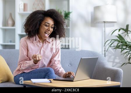 Donna eccitata con capelli ricci che celebra il successo mentre lavora su un notebook a casa. Emozioni felici, freelance o studente che raggiunge l'obiettivo, seduti su un divano grigio nel salotto moderno. Foto Stock
