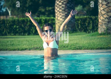 Una donna è in piedi in una piscina scintillante, con le braccia sollevate in un gesto di pura gioia. La luce del sole proietta un caldo bagliore sul suo viso sorridente mentre emana Foto Stock