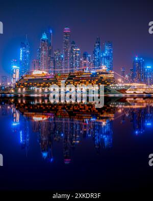 Una grande nave da crociera è attraccata nella Marina di Dubai di notte, con lo skyline della città e i suoi riflessi nell'acqua visibili sullo sfondo. Foto Stock
