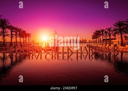 Un grande cartello per il Dubai Creek Harbour si trova sull'acqua al tramonto, con palme e uno skyline della città sullo sfondo. Foto Stock