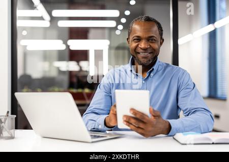 Un uomo d'affari professionista sorride con fiducia mentre utilizza tablet e notebook in un ufficio moderno e luminoso. Concetto di lavoro, tecnologia, successo e produttività in un ambiente professionale. Foto Stock