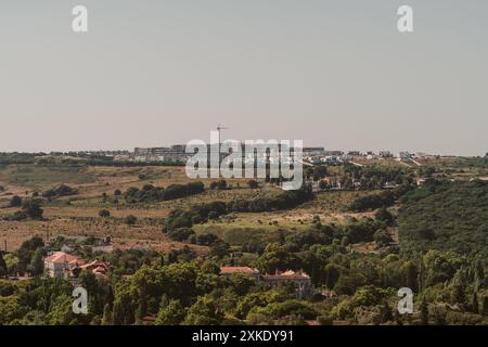 Una vista panoramica di un paesaggio collinare con case sparse ed edifici in costruzione all'orizzonte. Alberi verdi e vegetazione coprono la prua Foto Stock