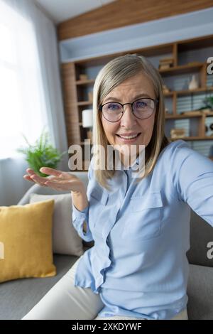 Donna anziana che indossa occhiali seduti sul divano e impegnata in videochiamata. Camera luminosa con cuscino giallo, ripiani in legno e piante verdi. Concetto di comunicazione Foto Stock