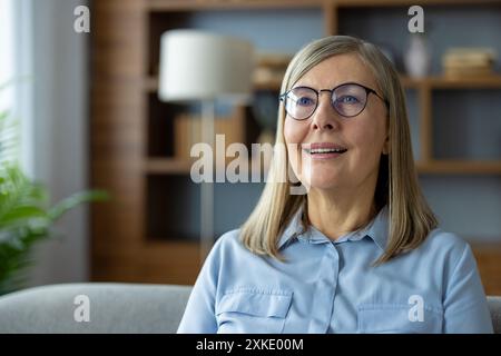 Donna matura sorridente con occhiali che sembrano felici mentre si siede in un accogliente soggiorno. Interni moderni con libreria e lampada sullo sfondo Foto Stock