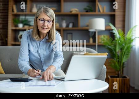 Donna anziana che lavora da casa con un laptop, che esamina documenti e utilizza la calcolatrice nel salotto moderno. Configurazione dell'ufficio domestico con scrivania e libreria in background. Concetto di produttività del lavoro remoto. Foto Stock