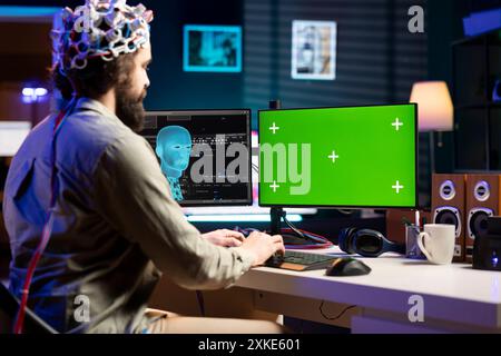 Tecnico IT che utilizza il dispositivo auricolare EEG, traduce i pensieri in comandi sul display del PC con schermo verde sulla scrivania. Amministratore di sistema che controlla le funzioni chiave del computer Chroma utilizzando Mind Foto Stock
