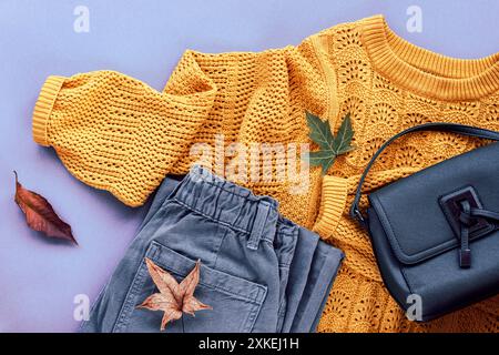 Giacenza piatta di maglione giallo donna, jeans grigi, borsa e foglie autunnali. Concetto di moda autunnale. Vista dall'alto, disposizione piatta. Foto Stock