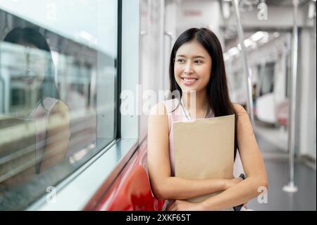 Una giovane dipendente asiatica, affascinante e positiva, sta prendendo un treno sopraelevato per andare al lavoro, seduto a sedere e guardando la vista dall'esterno. tran. pubblico Foto Stock