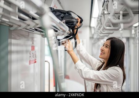 Una giovane ragazza asiatica felice e affascinante mette il suo zaino in un vano bagagli sopraelevato mentre si sposta su un treno sopraelevato o in metropolitana in una Foto Stock