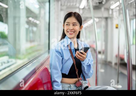 Una bella giovane donna asiatica è seduta su un treno sopraelevato con uno smartphone in mano, guardando la vista all'esterno, passando per il tragitto Foto Stock