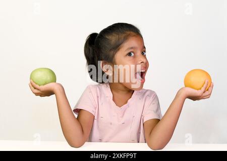 Una giovane ragazza lotta per scegliere tra una mela verde e un'arancia, la sua sorpresa e l'indecisione evidenti sul suo viso. Foto Stock