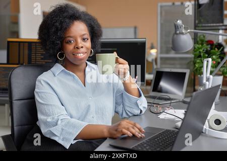 Ritratto di una giovane donna nera sorridente che lavora nell'ufficio di sviluppo IT seduto alla scrivania mentre prende una tazza di caffè guardando la fotocamera, copiando spazio Foto Stock