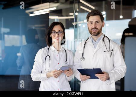 Ritratto di due medici in piedi insieme in ospedale. Sia indossando camici bianchi che stetoscopi. Medico donna che tiene la compressa mentre medico maschio tiene gli appunti. Foto Stock