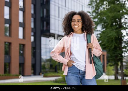 Giovane studente fiducioso con zaino in piedi davanti ad un edificio moderno. Sorridente e informale, trasuda energia positiva. Rappresentazione perfetta della vita studentesca, dell'istruzione e del successo in ambiente urbano Foto Stock