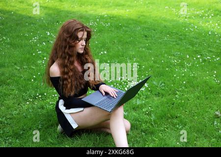 Bella ragazza con lunghi capelli di zenzero seduta con un computer portatile su un'erba nel parco estivo che lavora all'aperto Foto Stock