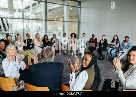 Un gruppo eterogeneo di persone applaude durante una sessione di workshop in un ambiente di ufficio contemporaneo, favorendo il lavoro di squadra e la collaborazione. Foto Stock