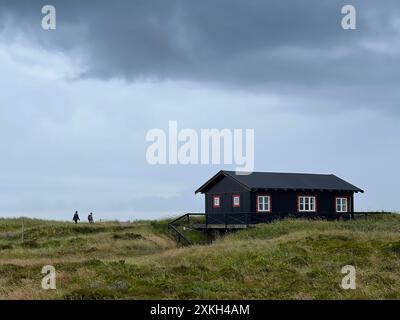 Danimarca, Skagen - 8 luglio 2024: Due turisti che si recano in una casa solitaria e pittoresca in stile danese su un prato sotto il cielo nuvoloso e scuro prima di piovere Foto Stock