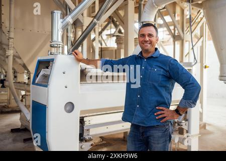 Un giovane uomo sorridente in abiti casual lavora all'ascensore. L'attraente agronomo regola le attrezzature. Il tecnico addetto alla lavorazione della granella controlla la smerigliatrice granella. Oper Foto Stock