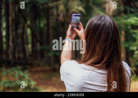 Una giovane donna in abiti casual scatta foto di paesaggi sul suo telefono. Vista dal retro. La turista scatta foto della foresta sul suo cellulare. Gli escursionisti amano il natu Foto Stock