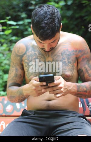 Un giovane uomo tatuato e in forma siede su una panchina della città, fissando intensamente il suo telefono. Il suo look rilassato ed elegante completa l'ambiente urbano Foto Stock