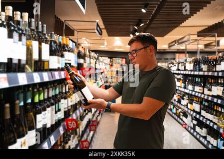 Un uomo in un corridoio di un negozio, che esamina attentamente una bottiglia di vino tra una varietà di opzioni. Consumatore maschile, bevendo sobrio Foto Stock