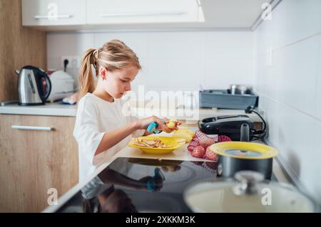 La giovane ragazza si concentra sulla preparazione del piatto di patate in una cucina moderna ed elegante. Pulisce la patata con un coltello per peeling atmosfera accogliente, mettendo in risalto la gioia Foto Stock