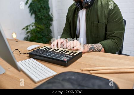Un giovane asiatico che indossa le cuffie si siede a una scrivania nel suo studio, suonando una tastiera MIDI mentre compone musica. Foto Stock