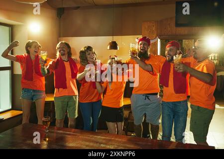 Amici, tutti in camicie arancioni e sciarpe rosse, bicchieri allegri, canto di gioia e felicità, celebrando la vittoria della squadra preferita al bar. Foto Stock