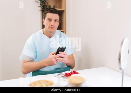 Ritratto di paziente maschio in cappotto medico utilizzando il telefono, riposo, mangiare, guadagnando forza prima di lunga operazione in clinica. Foto Stock