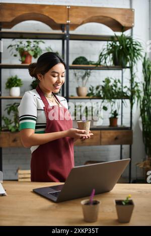 Una donna asiatica sorridente in un grembiule tiene una piccola pianta in vaso nel suo negozio di piante. Foto Stock