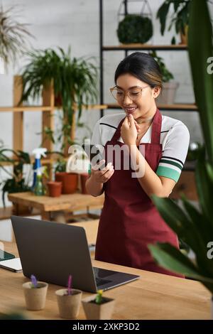 Una giovane donna asiatica, proprietaria di un negozio di piante, sorride mentre controlla il suo telefono in un momento di riflessione in mezzo al verde. Foto Stock