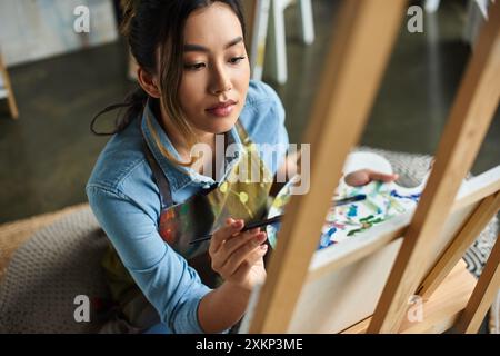 Una giovane artista asiatica, che indossa un grembiule, dipinge su una tela nel suo laboratorio. Foto Stock