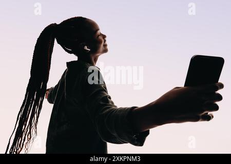 Silhouette di una giovane donna con capelli intrecciati che reggono uno smartphone e indossano auricolari. Si muove con grazia con tranquillità e piacere mentre ascolta Foto Stock