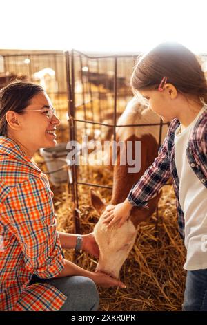 La giovane madre sorridente e la sua piccola figlia accarezzano un grazioso vitello in un capanno. Donna che ride in abiti casual e suo figlio si prendono cura degli animali domestici su fa Foto Stock