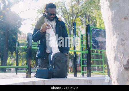 Black Man nel parco in piedi prendendo un asciugamano sudore dallo zaino, si sta preparando ad allenare i calisteni. Ha il suo zaino appoggiato su una panchina di cemento Foto Stock