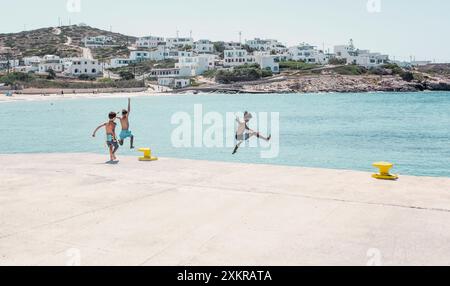 Tre ragazzi si tuffano nelle acque cristalline del Mar Egeo, con un pittoresco villaggio greco che si affaccia sulla scena. Foto Stock