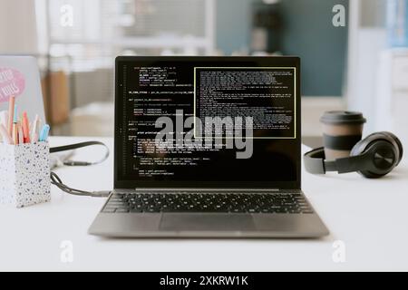 Primo piano medio di cuffie nere, forniture per ufficio e tazza di caffè accanto a un laptop argento con programma lanciato su un tavolo in legno bianco Foto Stock