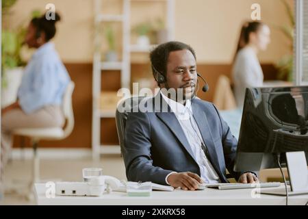 Operatore di call center afroamericano vestito con una tuta grigia che effettua videochiamate via computer mentre si trova in ufficio, circondato dai suoi colleghi birazziali Foto Stock