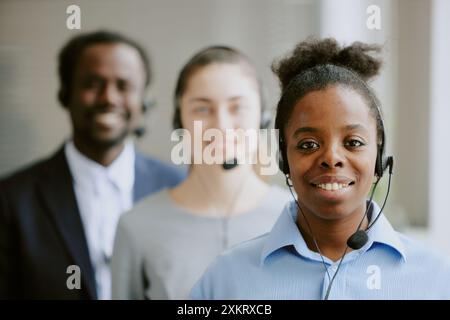 Ritratto di una giovane lavoratrice afroamericana che indossa la cuffia sorride ampiamente e guarda la fotocamera mentre la sua collega birazziale posa in sfocata Foto Stock
