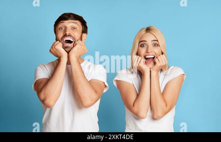 Un uomo e una donna sono mostrati sorridenti eccitati con le mani sulle guance. Entrambi indossano camicie bianche e stanno in piedi davanti a un bac blu Foto Stock